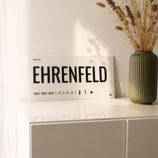Designschild „Ehrenfeld“ auf einem hellen Regal. Detailaufnahme des Acrylglas-Designschilds „Ehrenfeld“ von DasIstColonia mit minimalistischer Typografie und warmem Tageslicht. Design Straßenschild für Köln-Liebhaber