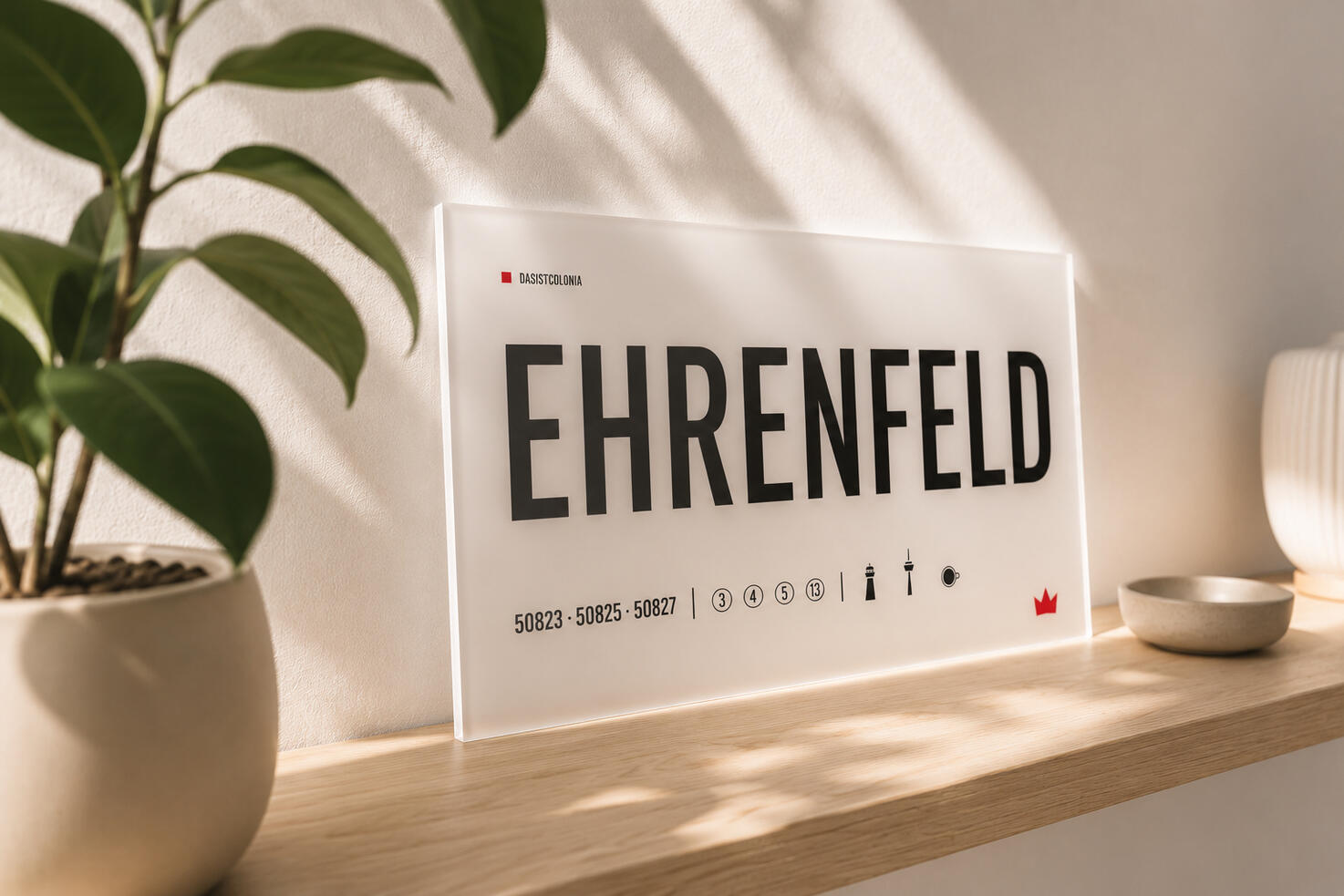 Designschild aus opalem Acrylglas „Ehrenfeld“ von DasIstColonia auf einem hellen Regal im warmen Tageslicht. Design Straßenschild für Köln-Liebhaber